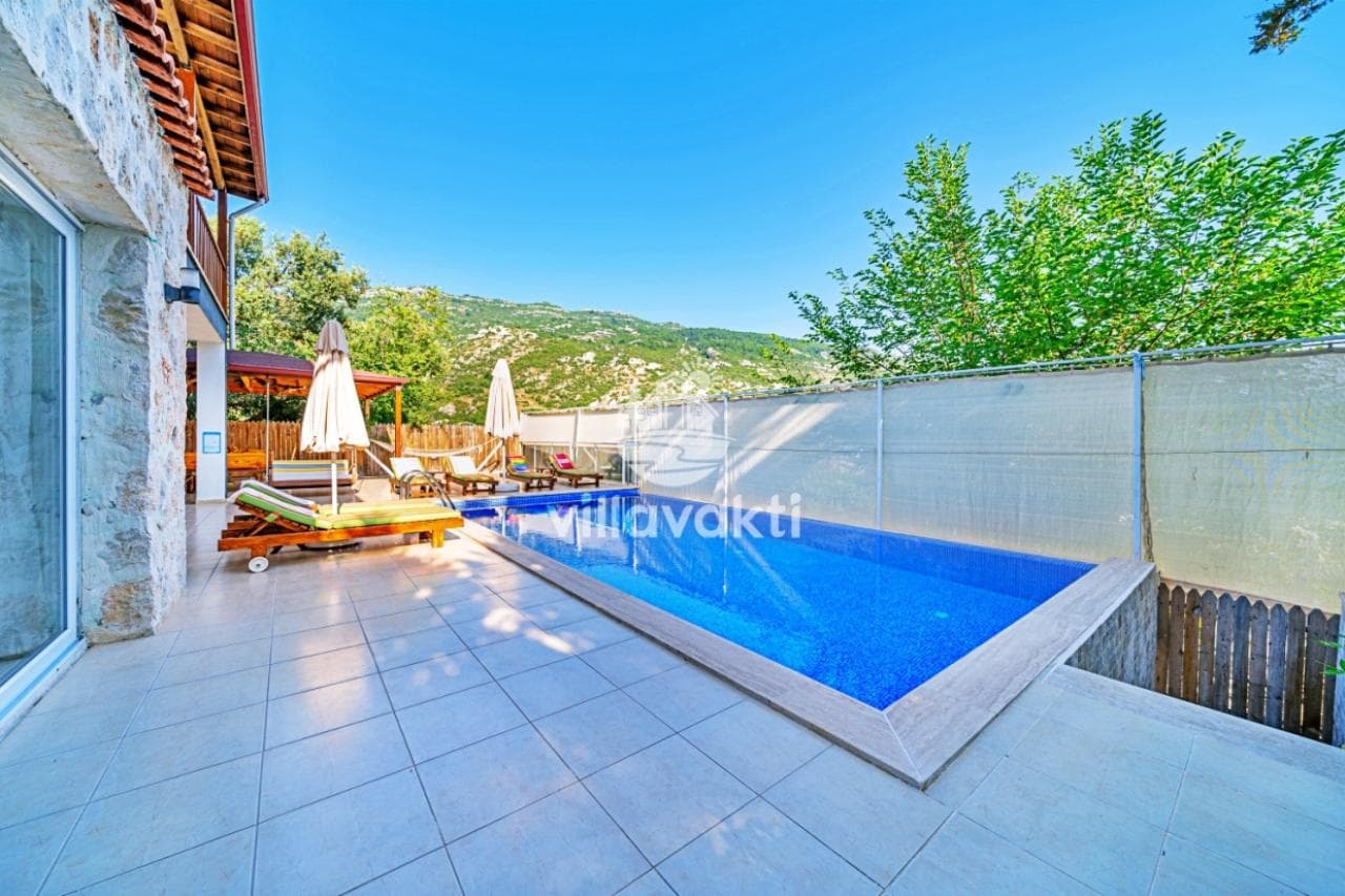 Taş duvarlı villanın terasında yüzme havuzu, şezlonglar ve ahşap pergola.